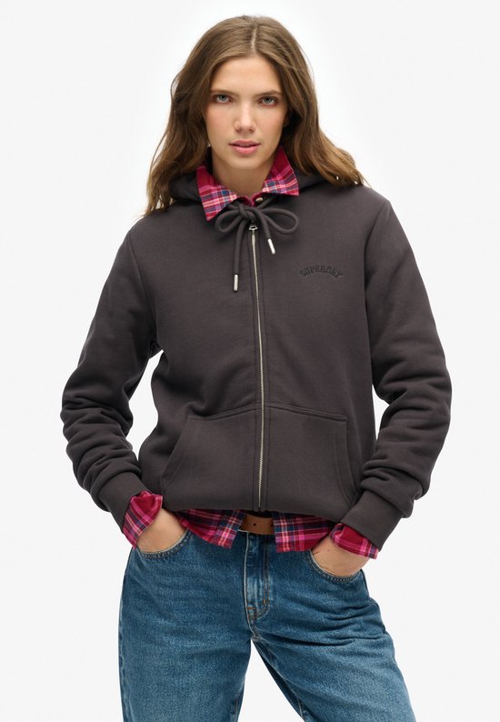 Model draagt een donkergrijs vest met rits over een rood-roze geruit overhemd en blauwe jeans.