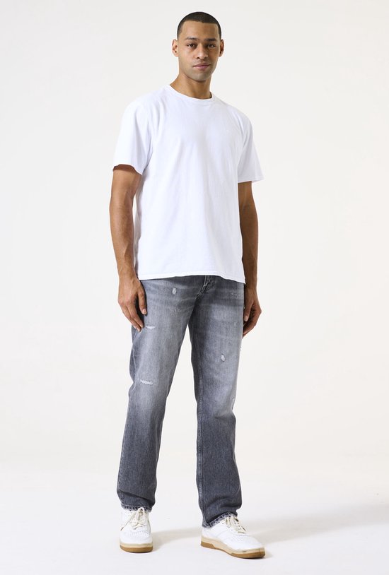 Man in een witte T-shirt en grijze jeans, met gescheurde details.