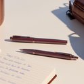 Deux stylos bordeaux sont posés sur un carnet ouvert. Une bouteille couleur cuivre est partiellement visible.