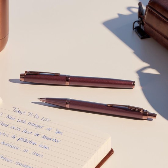 Deux stylos bordeaux sont posés sur un carnet ouvert. Une bouteille couleur cuivre est partiellement visible.