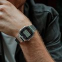 Close-up van de pols van een man met een zilverkleurig Casio horloge met metalen band, met een grijze denim jas op de achtergrond.