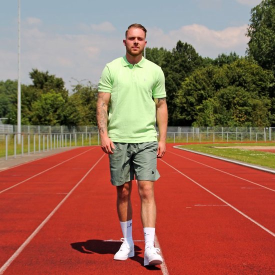 Man met tatoeages op een atletiekbaan, hij draagt een lichtgroen poloshirt en een grijze short.