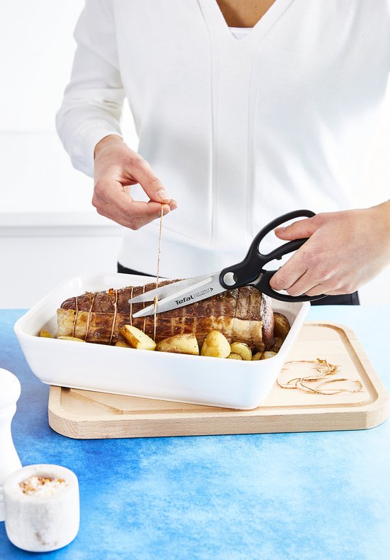 Zwarte keukenschaar snijdt touw van een gebraden rollade met aardappelen in een ovenschaal.