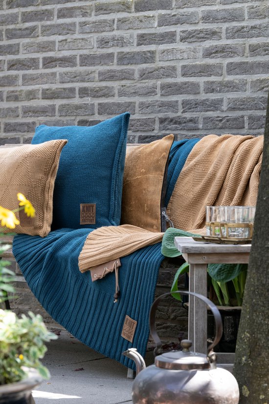 Een tuinbank met blauwe en bruine gebreide kussens en plaids. Een koperen theepot staat op een bijzettafel.
