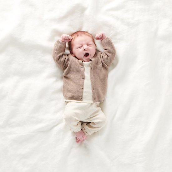 Baby in een beige gebreide cardigan, wit T-shirt en crème broek, liggend op wit laken.