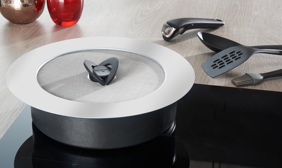 Zilverkleurig Tefal anti-spat deksel met een zwart hartvormig handvat, geplaatst op een pan op een inductiekookplaat.