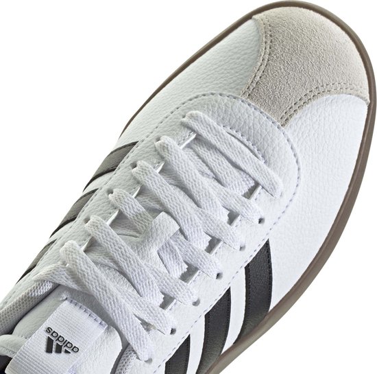 Detailopname van de witte Adidas sneaker: veters, grijze suède neuskap en zwarte strepen.