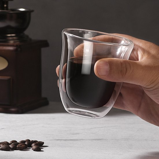 Verre à double paroi rempli de liquide sombre, tenu par une main. Grains de café au premier plan.