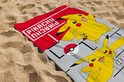 Een rode en grijze strandlaken met twee vrolijke Pikachu's op het zand. Bovenaan staat "PIKACHU" in witte letters. Onder de onderste Pikachu staat "#025".