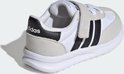 Witte kindersneaker met grijze suède overlays, zwarte Adidas-strepen en klittenbandsluiting.