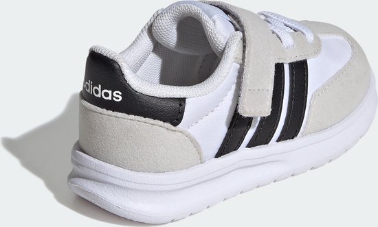 Witte kindersneaker met grijze suède overlays, zwarte Adidas-strepen en klittenbandsluiting.