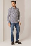 Man met een blauw-wit met bruine print overhemd en spijkerbroek, staand met hand in zak.