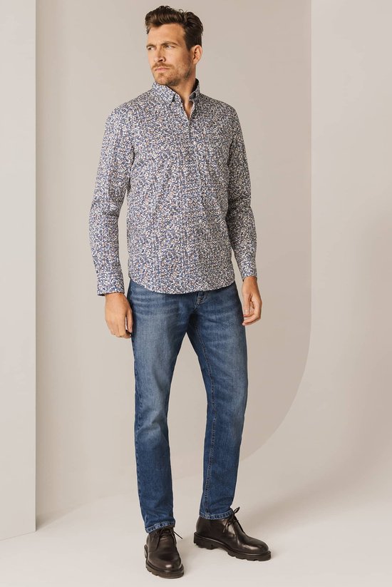 Man met een blauw-wit met bruine print overhemd en spijkerbroek, staand met hand in zak.