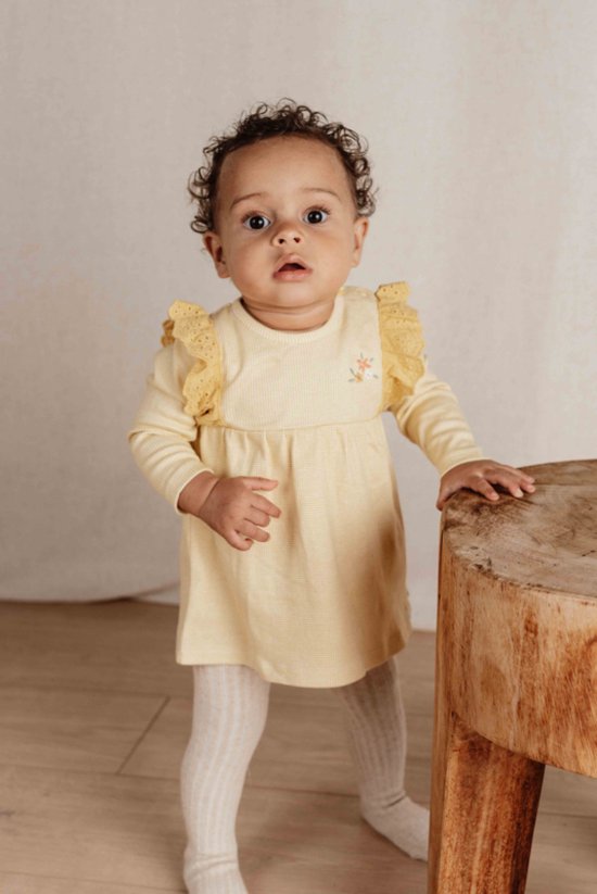 Baby in een geel geruit jurkje met ruches en bloemenapplicatie, wit gebreide maillot.