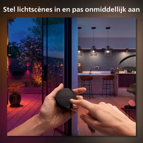 Een hand gebruikt de zwarte Philips Hue tap-dial switch om verlichting in een woonkamer en tuin te bedienen. De kamers hebben verschillende kleurenschema's.
