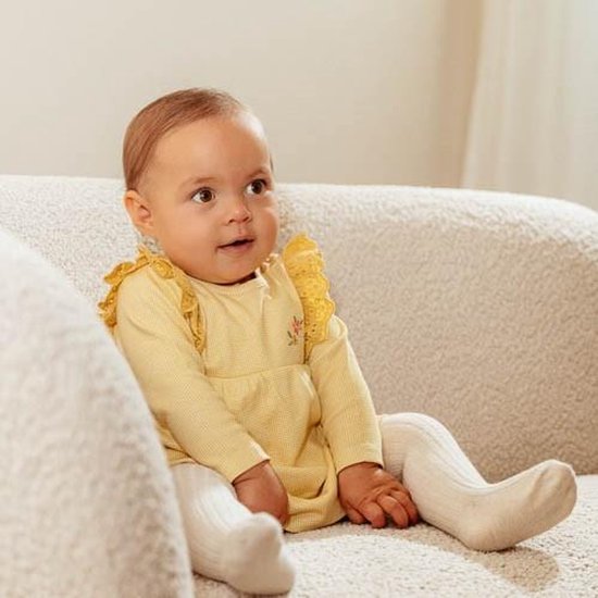 Baby in geel geruit jurkje met kanten kraagje en bloemenprintje op de borst, zittend op een bank.