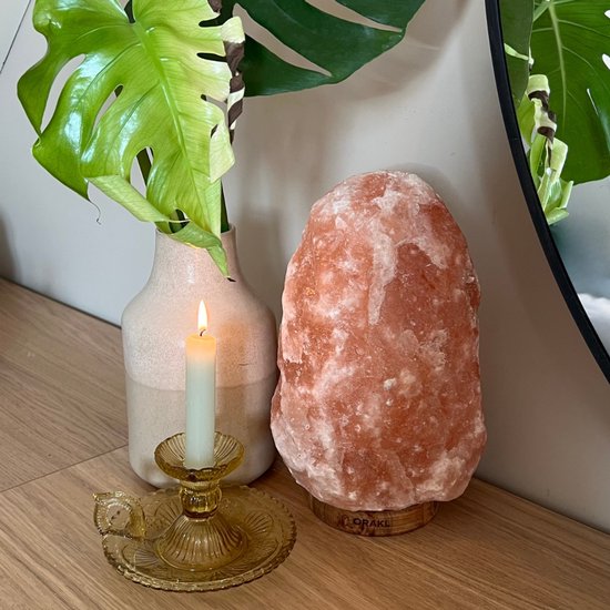 Roze Himalaya zoutlamp met houten voet, naast een vaas met Monstera blad en gele kaarsenstokhouder met brandende kaars.