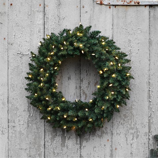 Donkergroene kerstkrans met warme witte lampjes, opgehangen aan een verweerde houten deur.