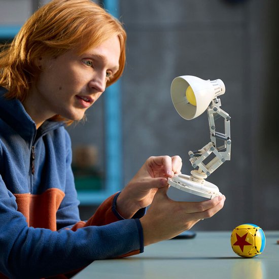 Persoon bouwt een wit LEGO Luxo Jr.-bureaulamptoestel naast een gele bal met een rode ster.