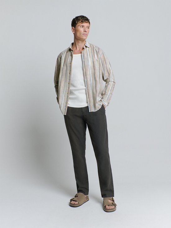 Model in een beige linnen mix blouse met verticale strepen, gedragen met een witte tanktop en donkere broek. De outfit wordt gecompleteerd met bruine sandalen.