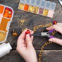 Handen maken sieraden met een tang en een doos met diverse kralen en onderdelen op een houten achtergrond.