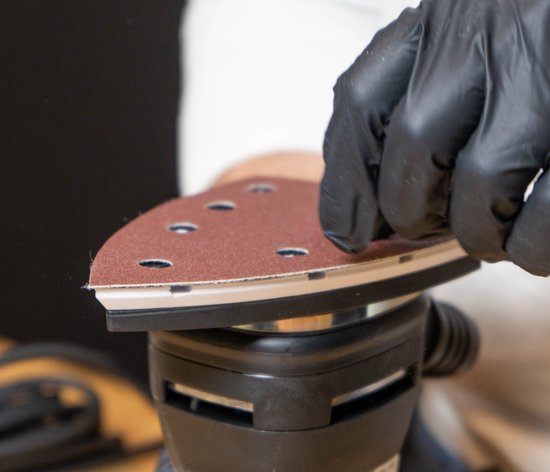 Een hand met zwarte handschoen plaatst bruin schuurpapier op een schuurmachine.
