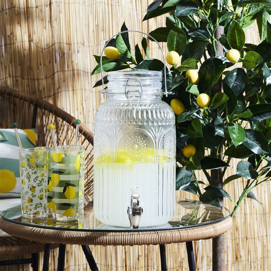Glazen drankdispenser met kraantje, gevuld met gele drank, naast glazen met citroenprint. Staat op een tafel onder een citroenboom.
