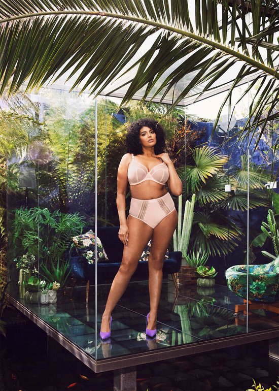 Model met krullend haar staat in een tropische setting, draagt een zachtroze beha en high-waist slip.