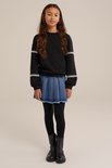 Meisje met lang bruin haar, draagt een zwarte sweater met witte banden op de mouwen, een spijkerrok en zwarte legging.