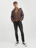Man met handen in zakken, draagt bruine pufferjas, bruine trui met rits en donkergrijze slim fit jeans.