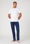 Man in een witte T-shirt en donkerblauwe Wrangler jeans, gedragen met bordeauxrode sneakers.