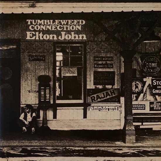 Elton John - Tumbleweed Connection album cover. Zwart-wit foto van een man die voor een winkel zit. De tekst 'Tumbleweed Connection' en 'Elton John' staat bovenaan.
