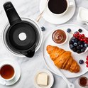 Glazen waterkoker naast een ontbijt met koffie, thee, croissant en fruit.