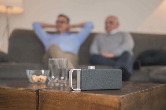 Zwarte luidspreker met zilveren handgreep op een houten salontafel. Twee mannen kijken tv op de achtergrond.