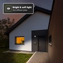 Zwarte wandlamp met sensor verlicht de entree van een moderne woning met witte gevel en zwarte accenten.