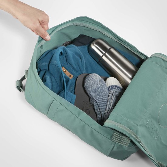 Open groene Kånken rugzak met daarin een thermosfles, kleding en sokken.