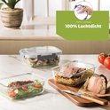 Glazen vershouddoos met vlees en rijst, naast een salade en vlees op een snijplank.