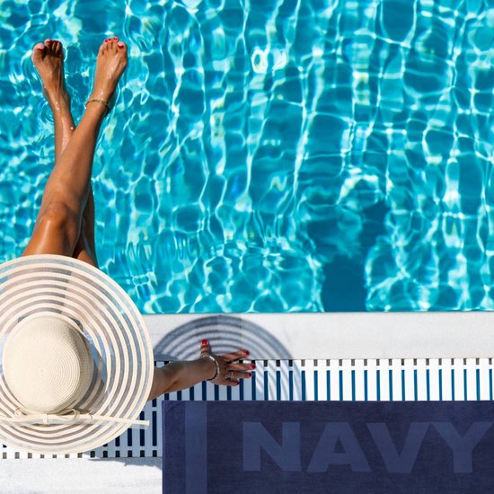 Persoon in zomerhoed en badkleding bij een zwembad met een marineblauw strandlaken met NAVY-opdruk.