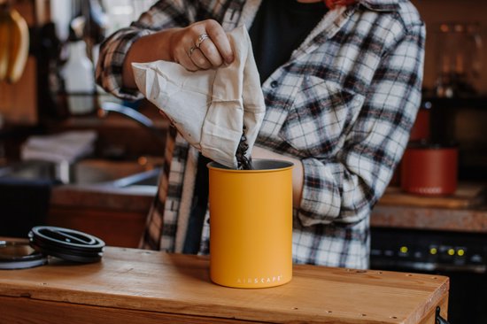 Handen met ringen gieten koffiebonen uit een zak in een matgele Airscape voorraadpot.