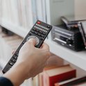 Hand houdt zwarte afstandsbediening vast voor een zwart micro-stereosysteem tussen boeken.