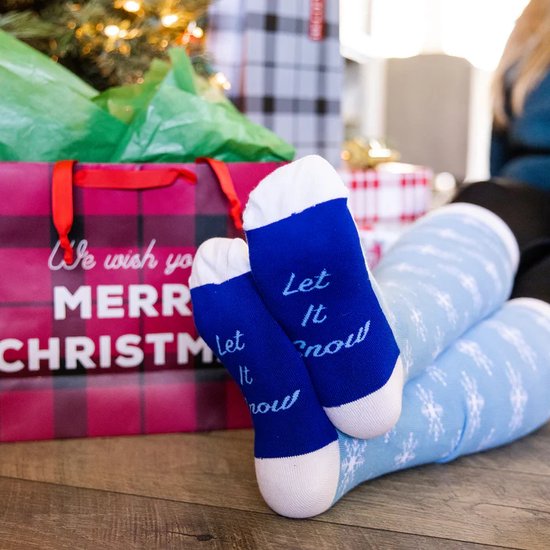 Blauwe kerstsokken met witte sneeuwvlokken-opdruk en tekst 'Let It Snow', gedragen door iemand bij een kerstboom en cadeaus.