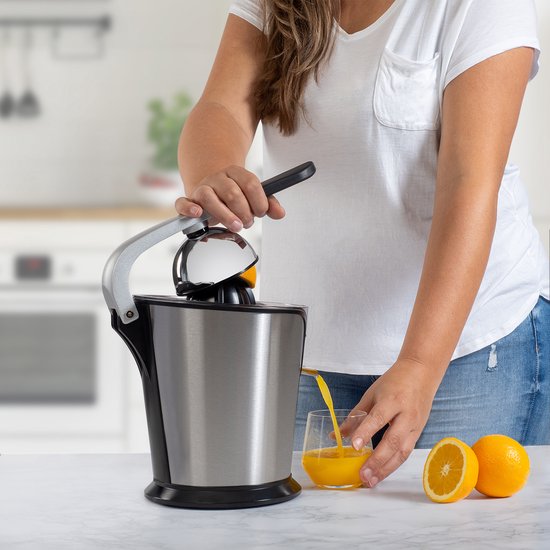 Vrouw gebruikt zilverkleurige KitchenBrothers citruspers met pershendel om oranje sap te persen in een glas. Twee sinaasappels liggen ernaast.