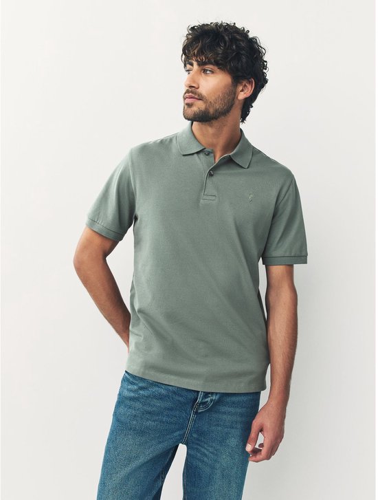 Man in een groen piqué poloshirt en spijkerbroek, leunend tegen een witte muur, kijkend naar de zijkant.