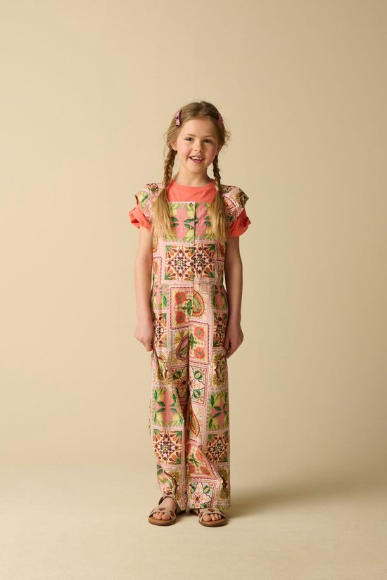 Meisje met blonde vlechten staat op een lichte achtergrond en draagt een lichtroze jumpsuit met een veelkleurig patchwork-patroon en oranje shirt met korte mouwen.