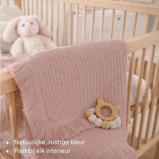 Lichtroze mousseline dekentje met wafelstructuur en franjes, liggend in een houten ledikant naast baby speelgoed.