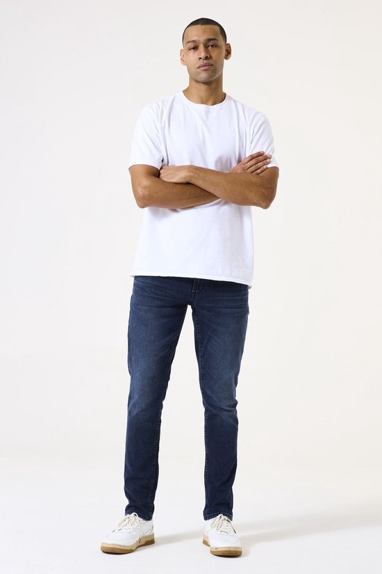 Man draagt een effen witte T-shirt met gekruiste armen en een donkerblauwe slim fit jeans.
