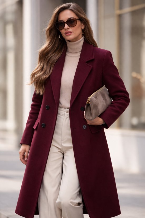 Vrouw in een bordeauxrode lange jas met reverskraag, beige coltrui, crème broek en zonnebril.