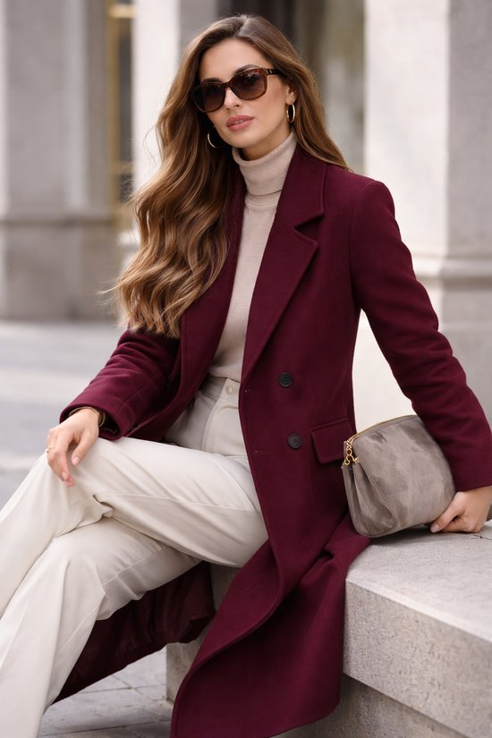 Vrouw zittend in een bordeauxrode lange jas met reverskraag, beige coltrui, crème broek en zonnebril.