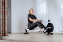 Een lachende vrouw met blonde paardenstaart zit op de VirtuFit roeimachine in een lichte ruimte.