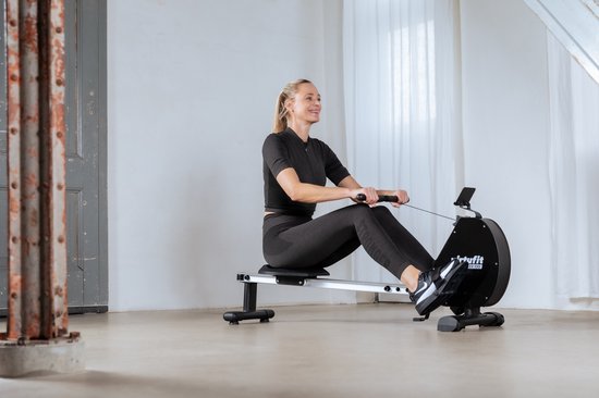 Een lachende vrouw met blonde paardenstaart zit op de VirtuFit roeimachine in een lichte ruimte.
